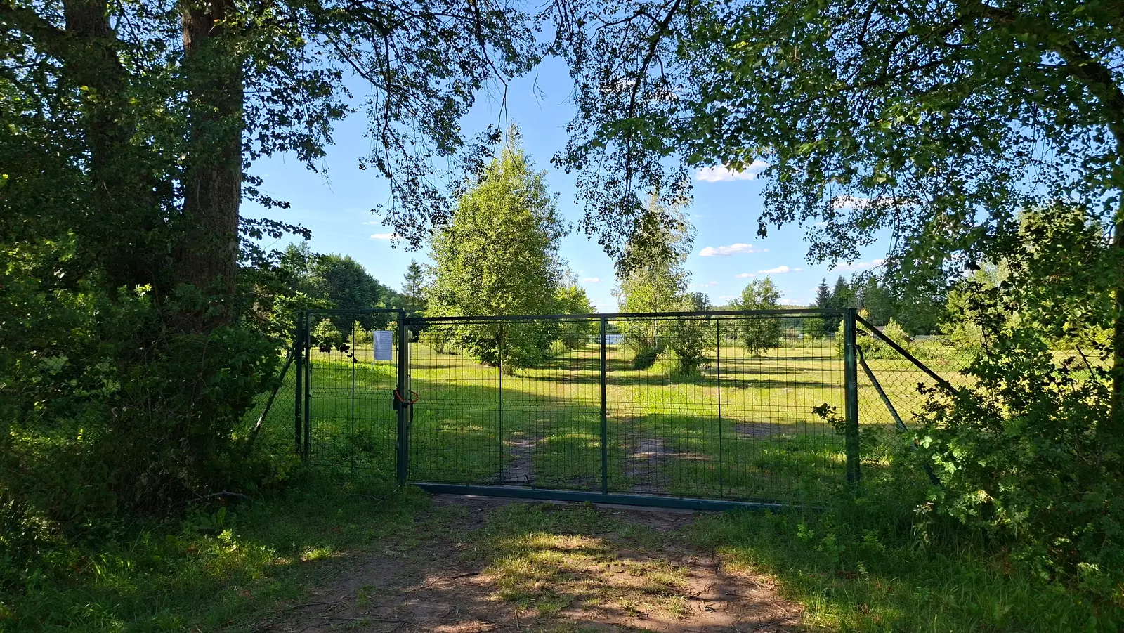 Zdjęcie o proporcjach 16:9 przedstawia zieloną, metalową bramę wjazdową umieszczoną na tle słonecznego, wiejskiego krajobrazu. Pierwszy plan zajmuje piaszczysta droga prowadząca do ogrodzenia z siatki, otoczonego przez gęste korony drzew rzucające cień na ziemię. Przez ażurowe skrzydła bramy rozpościera się widok na rozległy teren zielony z pojedynczymi drzewami i krzewami w pełnym słońcu. Na lewym skrzydle bramy znajduje się biała tabliczka informacyjna, na której widnieje napis: „TEREN PRYWATNY / WSTĘP WZBRONIONY / Właściciel nieruchomości”. Całość kompozycji zachowana jest w letniej, nasyconej kolorystyce, gdzie błękitne niebo z delikatnymi obłokami kontrastuje z soczystą zielenią roślinności.